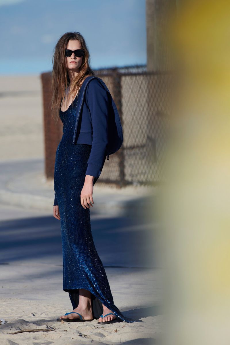 A brunette woman wearing a blue dress and jacket standing on a beach. She's wearing sunglasses and sandals in a fashion-style shot.