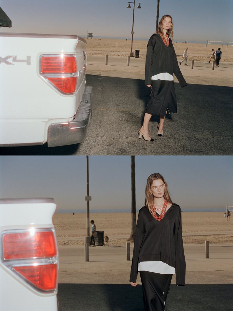 Fashion model posing on a street near a beach, wearing a black sweater, white shirt, and black skirt, with a white truck parked nearby.