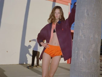 Model leaning against a palm tree wearing an oversized burgundy coat and bright orange shorts against an urban wall.