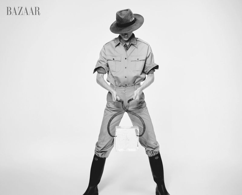 Person in overalls and hat holds ice block with metal tongs in a stark studio setting, black and white, high-fashion shot.