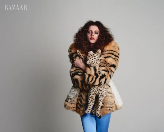 A woman with curly hair and unique makeup poses in a patterned fur coat holding a stuffed cheetah in a studio shot for fashion.