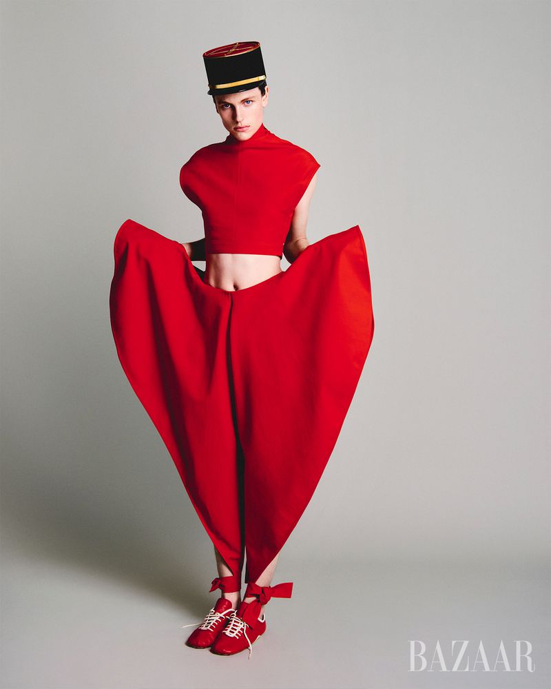 Fashion photo of a model in a red top, oversized pants, matching shoes, and a cap posing in a studio with a solid grey backdrop.
