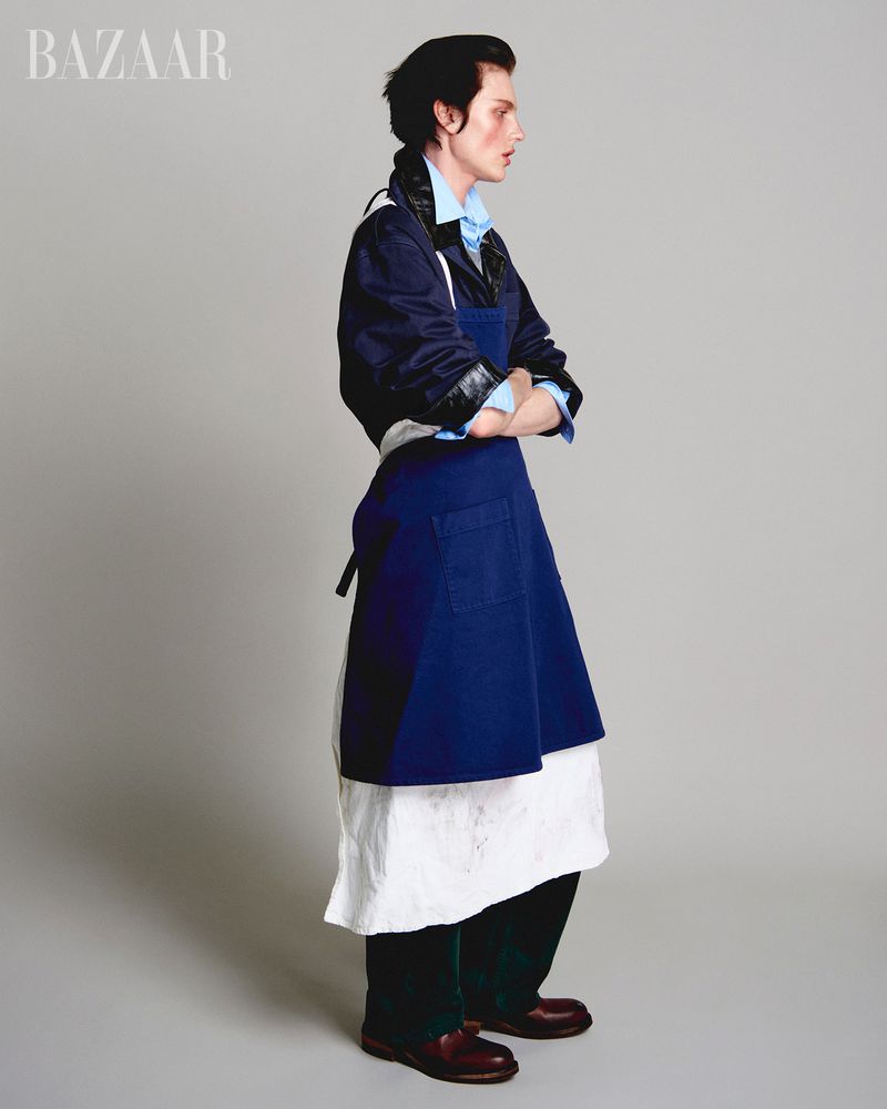 Fashion editorial shot of a person in a blue button-down shirt, navy jacket, and blue apron, standing in a photo studio with a grey backdrop.