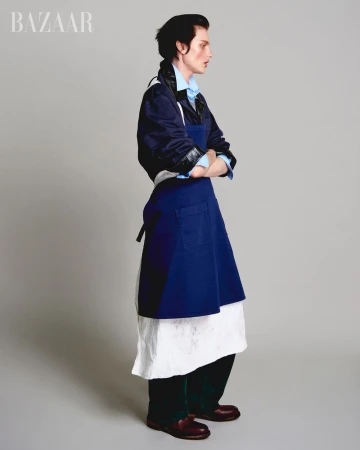 Fashion editorial shot of a person in a blue button-down shirt, navy jacket, and blue apron, standing in a photo studio with a grey backdrop.
