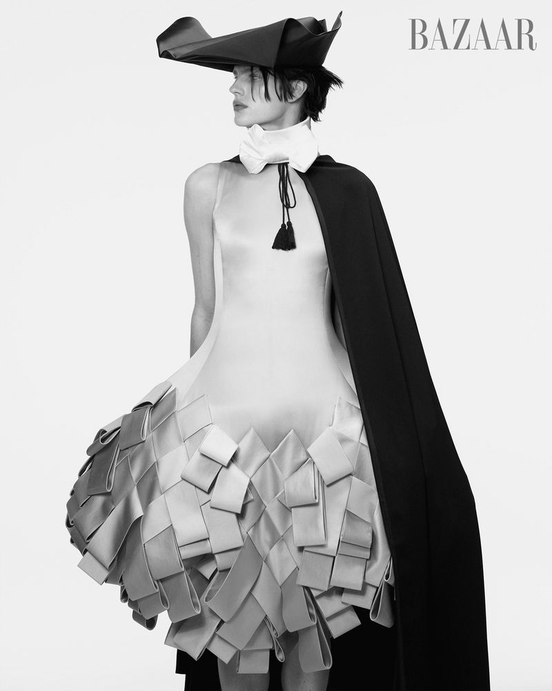 Black-and-white portrait of a woman in an avant-garde dress and tricorn hat, accessorized with a cape and high collar, in a studio setting.