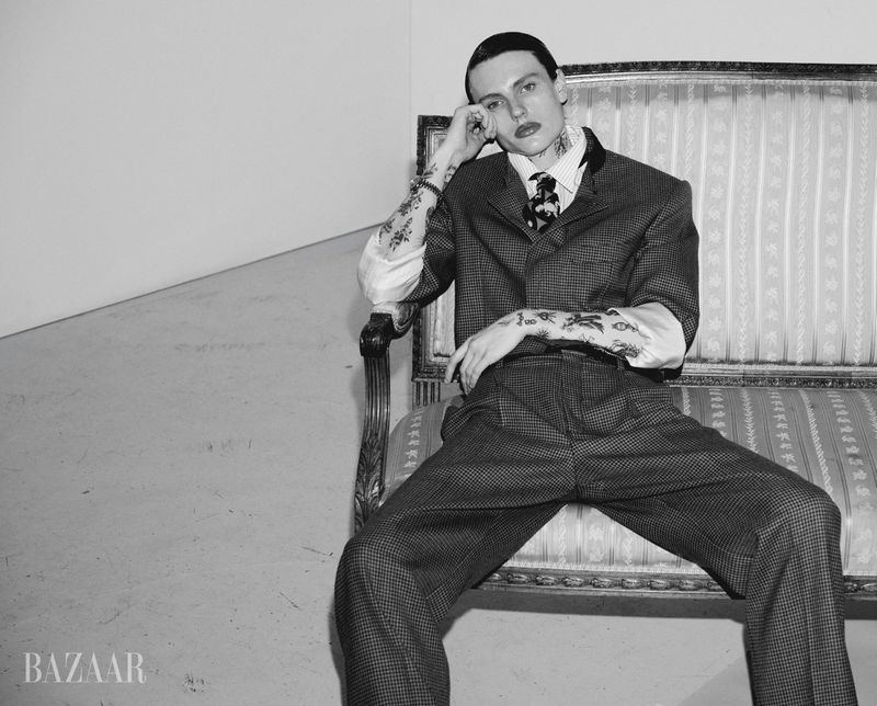 A fashionable adult male with tattoos poses on a patterned couch in a photo studio, wearing a checkered suit, in black and white.