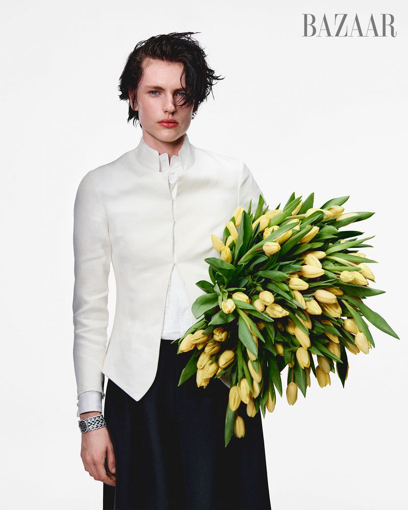 A woman with short black hair stands indoors, holding a bouquet of yellow tulips. She wears a white jacket and black skirt.