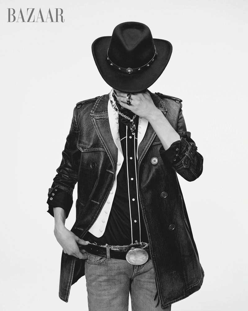Person wearing a cowboy hat, denim jacket, and jeans in a black and white studio portrait for Bazaar magazine with white backdrop.