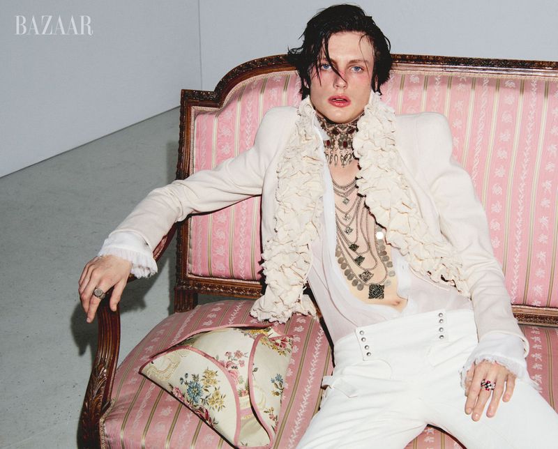 A person lounges on a pink couch, posing in a white jacket, matching pants, and multiple necklaces against a grey background.