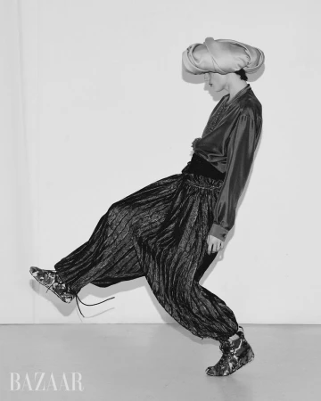 Editorial fashion shot: Person in a head wrap, shiny top, and patterned pants strikes a playful pose against a white backdrop. Bazaar logo bottom left.