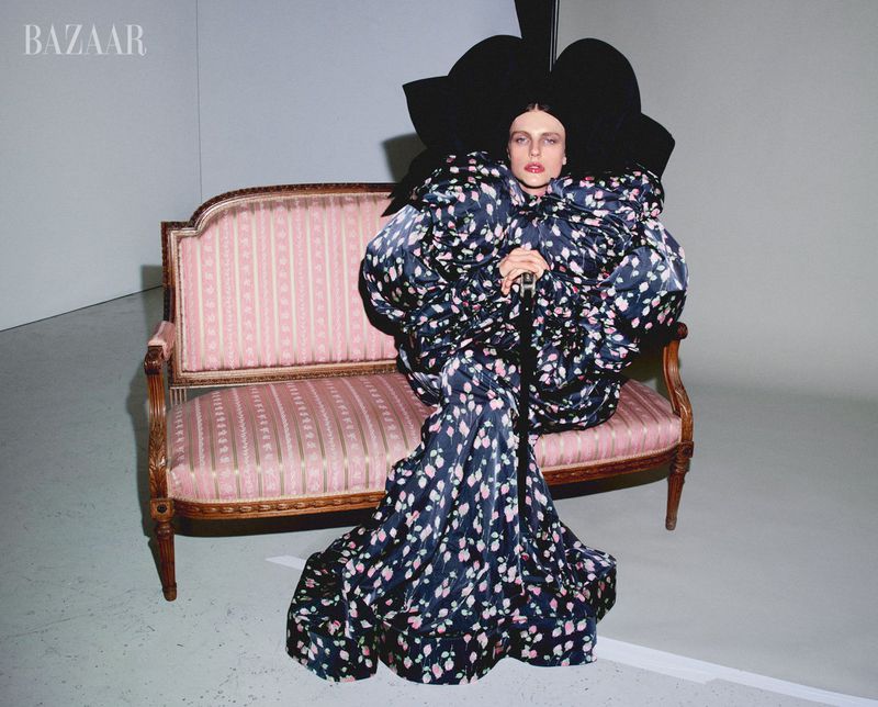 Fashion shot of a person in an elaborate floral dress and large hat sitting on a pink settee in a photo studio setting.