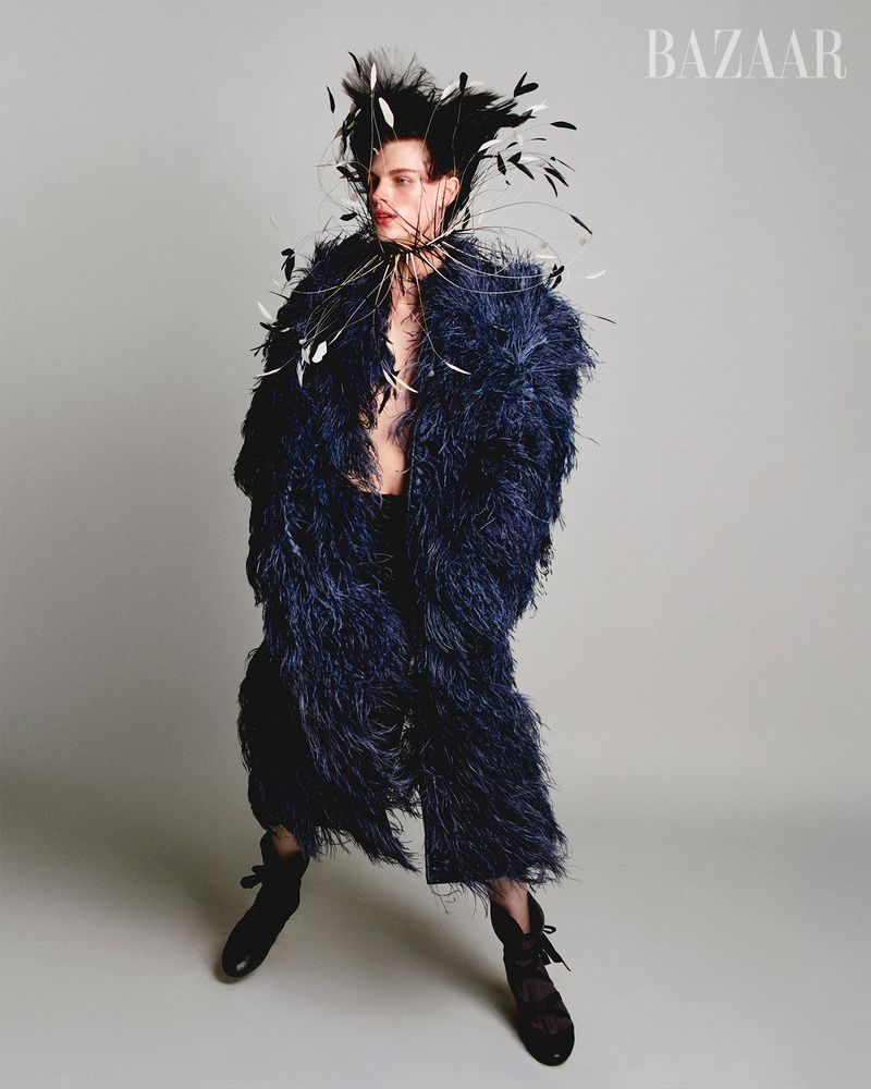 Person poses in a textured navy coat with a feathered headdress and black lace-up boots, set against a plain grey studio backdrop.