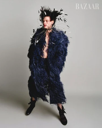 Person poses in a textured navy coat with a feathered headdress and black lace-up boots, set against a plain grey studio backdrop.
