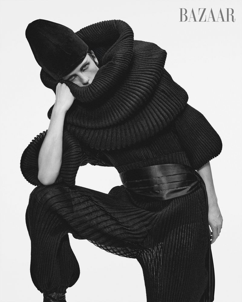 Fashion editorial shot of a person in a voluminous black outfit with textured fabric, posing against a bright background. Bazaar logo visible.