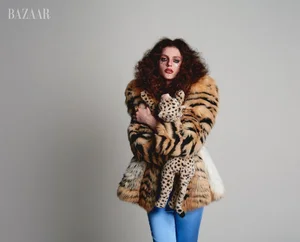 Model with voluminous curly hair in a tiger-print faux fur coat holding a stuffed cheetah against a neutral grey background