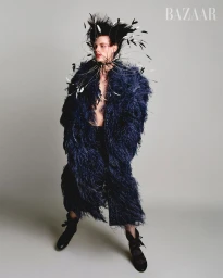 Model wearing a dark feather coat and elaborate feathered headpiece against a neutral grey studio background