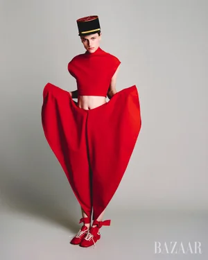 Model in red outfit holding out voluminous trousers, wearing a pillbox hat against a grey seamless background