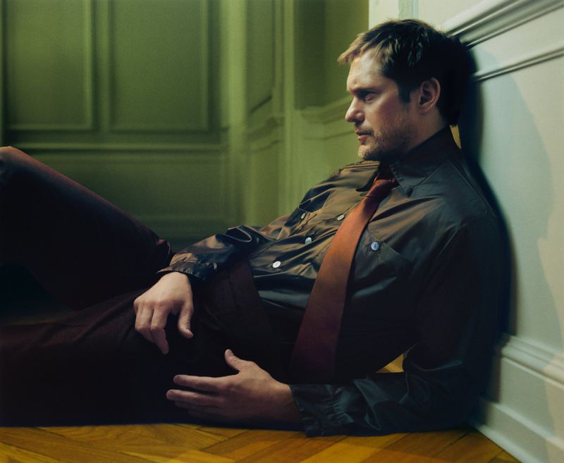 Portrait of a handsome man with a tie laying on a hardwood floor against a wall in an olive green paneled room in eye-level angle.
