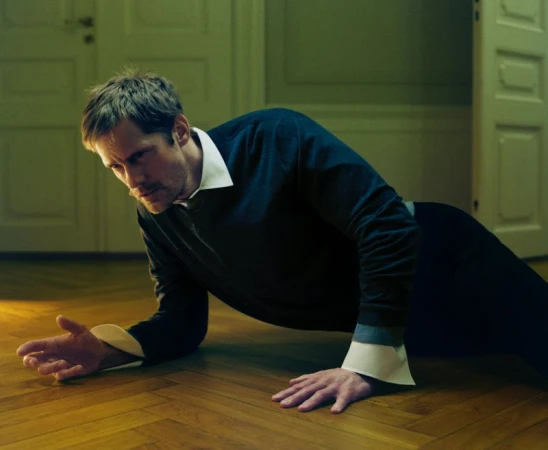 Man in a navy sweater does push-ups indoors. He is on a hardwood floor near a partially open white door, illuminated with soft lighting.