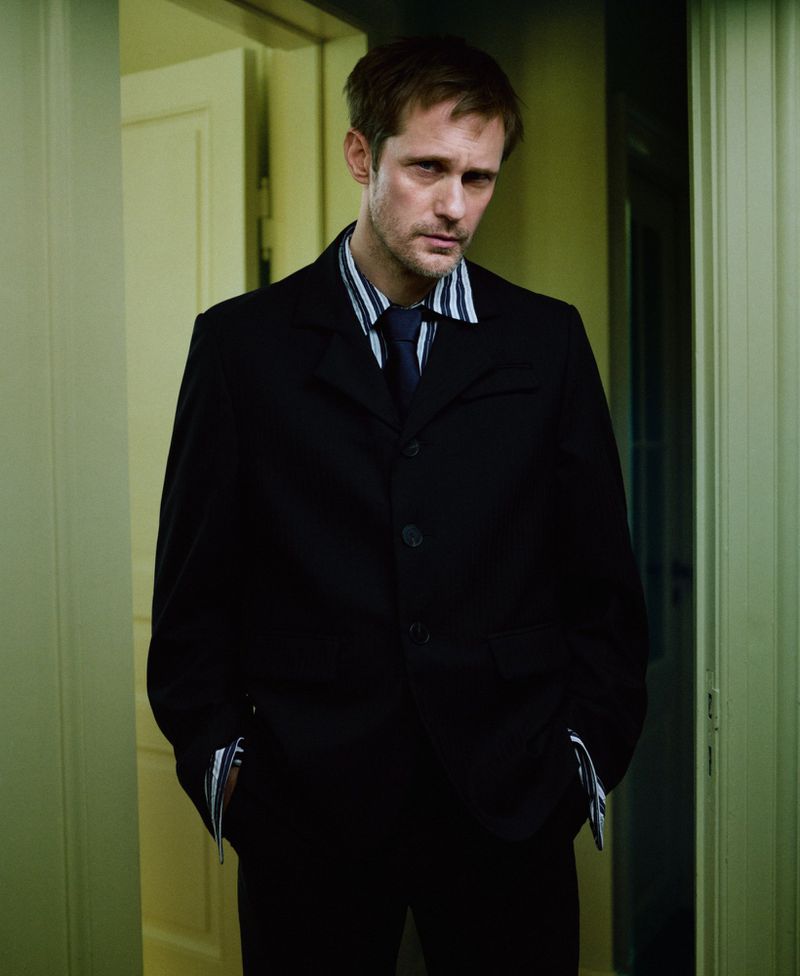Alexander Skarsgard wearing a dark suit and striped shirt standing indoors in a hallway for M Le magazine du Monde