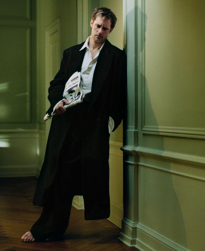 An adult male model leans against a wall indoors, holding a newspaper in one hand, wearing a black coat, white shirt, and dark pants.
