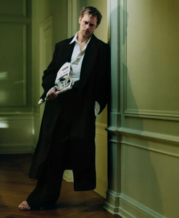 An adult male model leans against a wall indoors, holding a newspaper in one hand, wearing a black coat, white shirt, and dark pants.