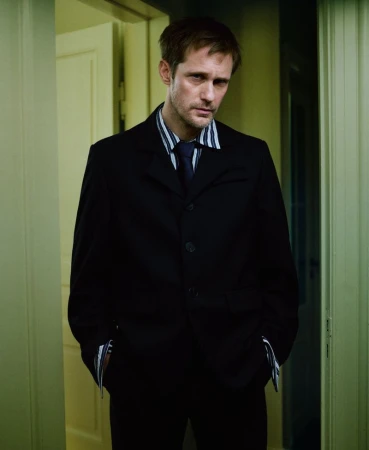 Alexander Skarsgard wearing a dark suit and striped shirt standing indoors in a hallway for M Le magazine du Monde