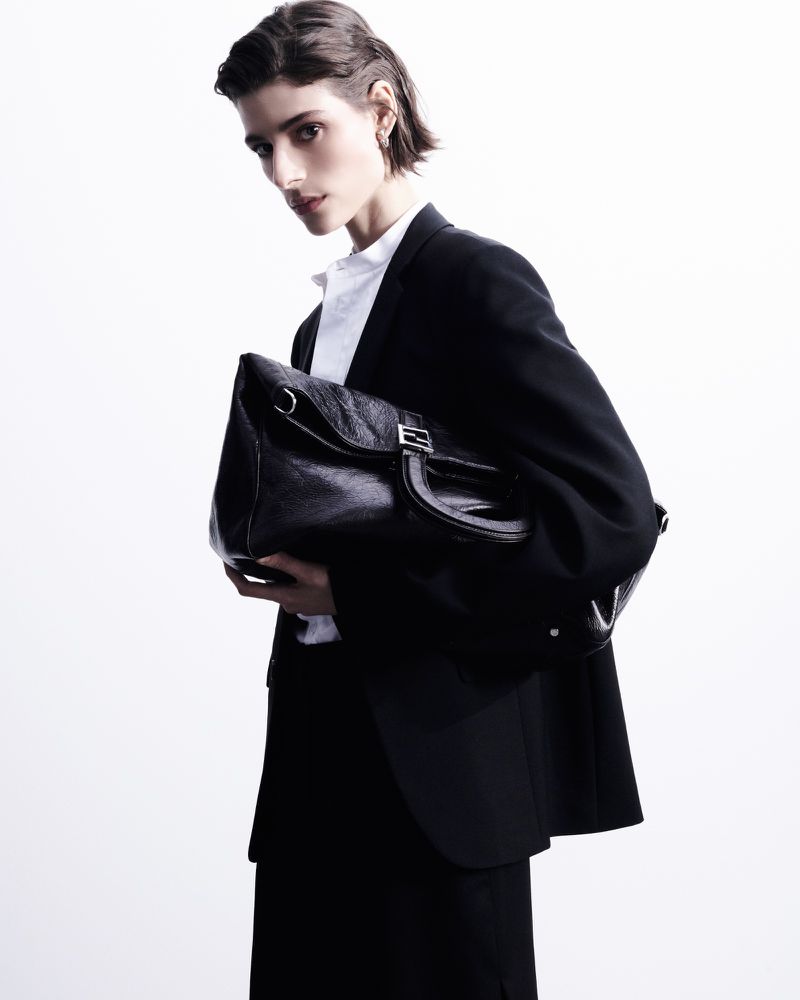 Fashionable woman in a black suit and white shirt holding a black leather satchel bag against a bright, neutral background in a studio setting.