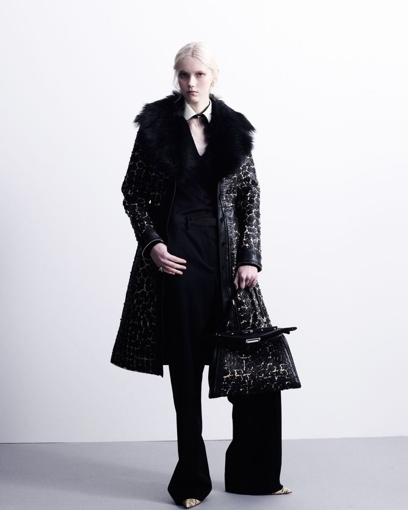 A woman with pale skin and blonde hair stands in a studio wearing a patterned black coat with a fur collar, black top and pants, and carrying a patterned purse.