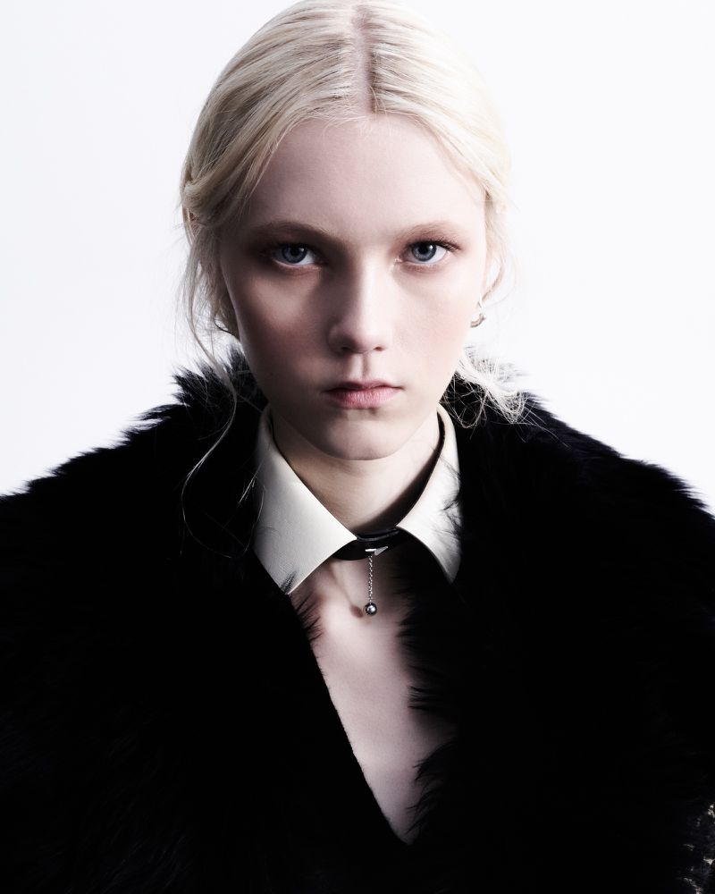 Portrait of a light-skinned female with blond hair, wearing a white shirt, black fur coat, and choker, against a bright white background.