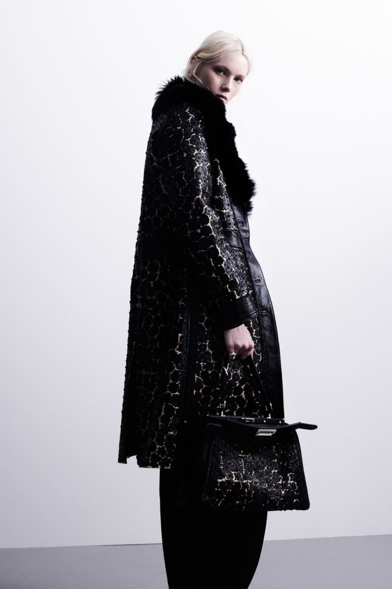 A woman in a black coat with fur collar and cuffs, holding a matching handbag, poses against a clean white studio backdrop.