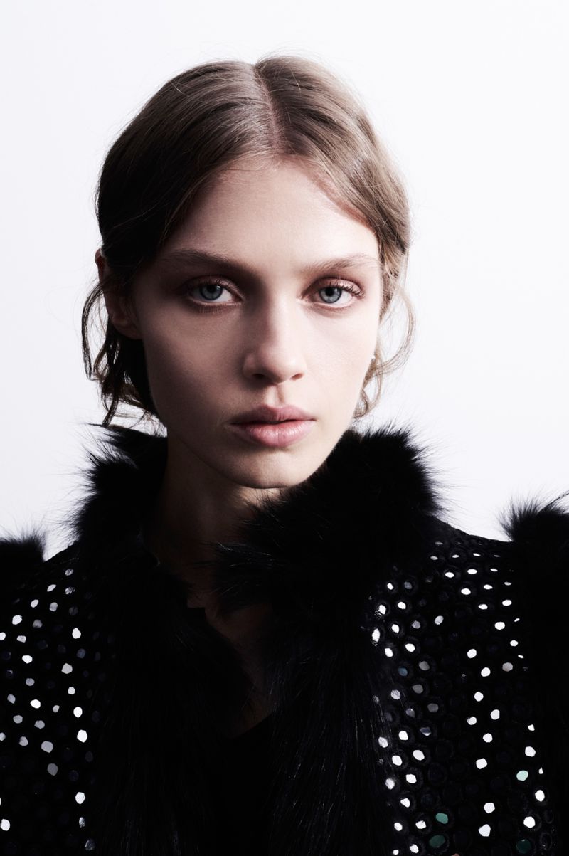 Close-up portrait of a woman with light brown hair and blue eyes wearing a black jacket with silver sequins on a white background.