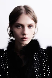 Portrait of a female model wearing a black embellished jacket with fur trim against a plain white background.