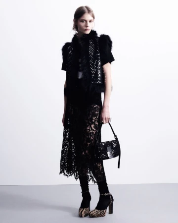 Model in black lace skirt and embellished vest holding a studded handbag against a plain white studio backdrop.