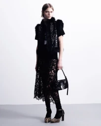 Model in black lace skirt and embellished vest holding a studded handbag against a plain white studio backdrop.