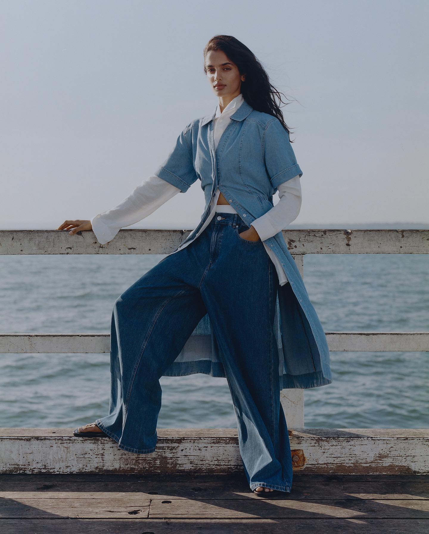 Brunette woman with blue denim shirt and wide-leg jeans poses on a pier with ocean and sky backdrop in an outdoor fashion shot.