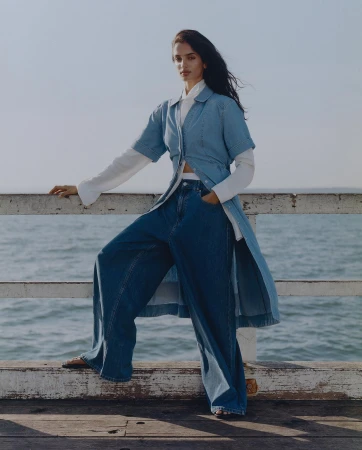 Brunette woman with blue denim shirt and wide-leg jeans poses on a pier with ocean and sky backdrop in an outdoor fashion shot.