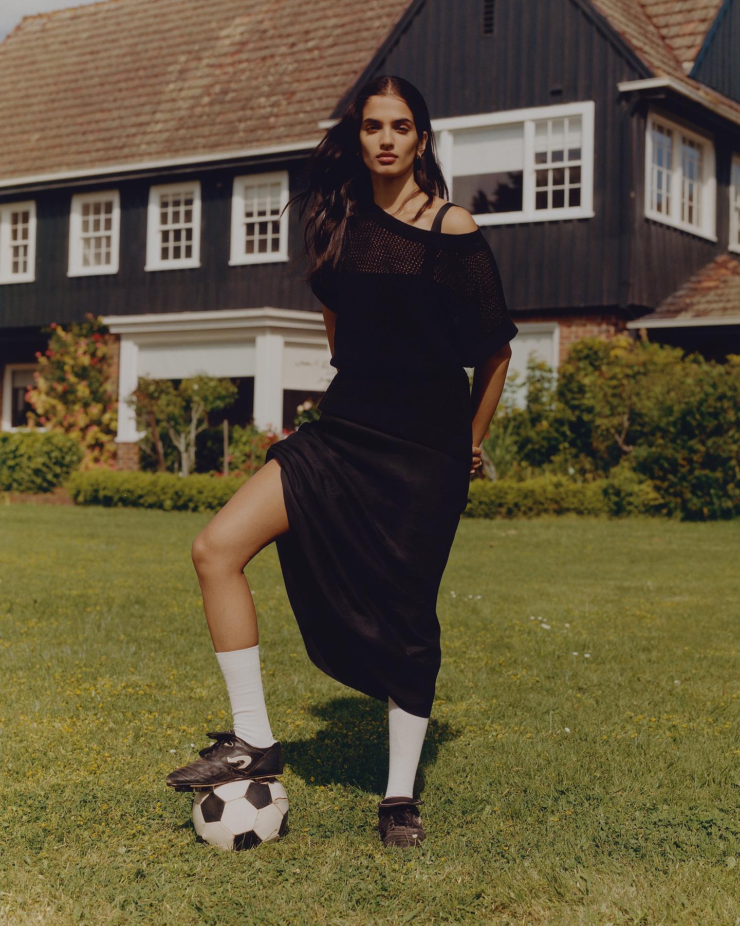 An adult woman poses outdoors in a black outfit, white socks, and soccer cleats, with her foot on a soccer ball in a yard.
