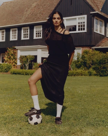 An adult woman poses outdoors in a black outfit, white socks, and soccer cleats, with her foot on a soccer ball in a yard.