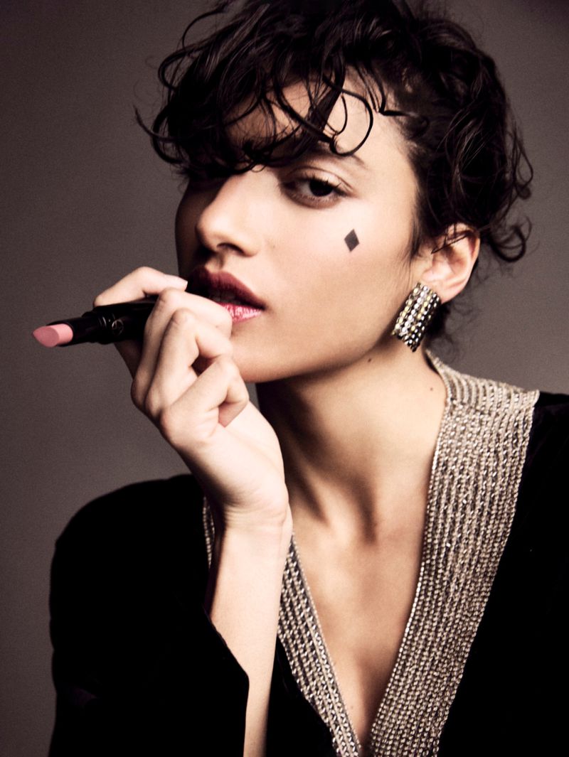 Close-up studio shot of an adult woman applying pink lipstick, wearing a black velvet top with beaded trim and sparkling earrings.