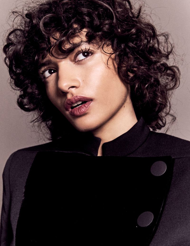 Close-up studio portrait of an adult woman with curly dark hair and a dark blazer-style jacket in front of a neutral background.