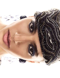 Close-up portrait of a model wearing a heavily beaded black cap and patterned garment against a bright white background
