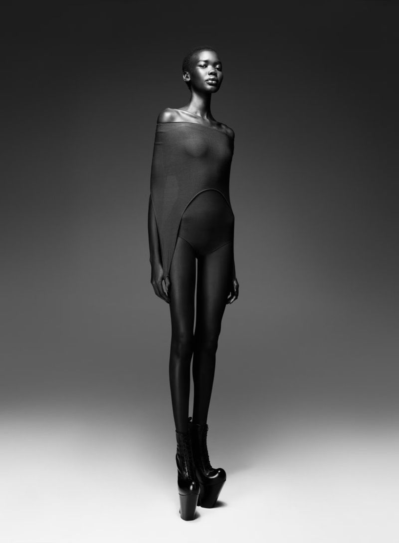 Black and white fashion shot of a slender, dark-skinned woman in a minimalist garment and platform boots, posing against a neutral gradient backdrop.