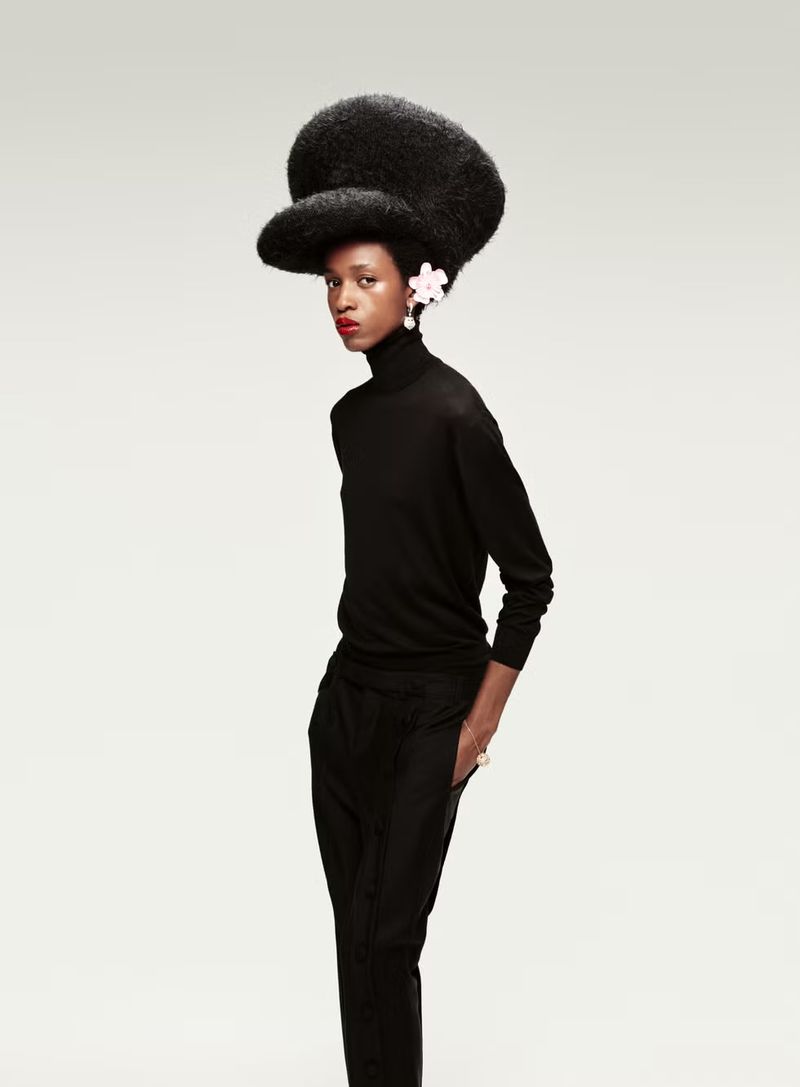 Woman in a black turtleneck and pants posing in a studio with afro hair and a flower hair clip. Fashion studio shot on white.