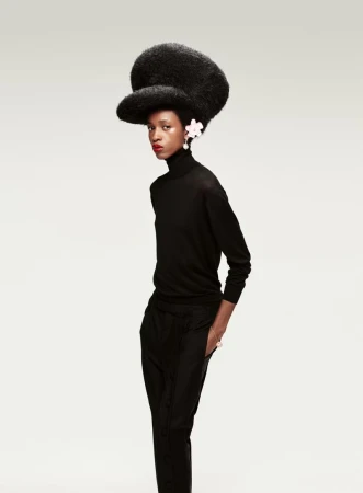 Woman in a black turtleneck and pants posing in a studio with afro hair and a flower hair clip. Fashion studio shot on white.