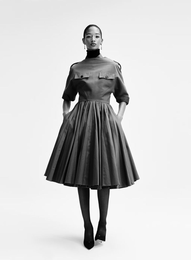 A woman stands centered against a white background, wearing a midi dress with a pleated skirt, epaulettes, and a dark turtleneck underneath.