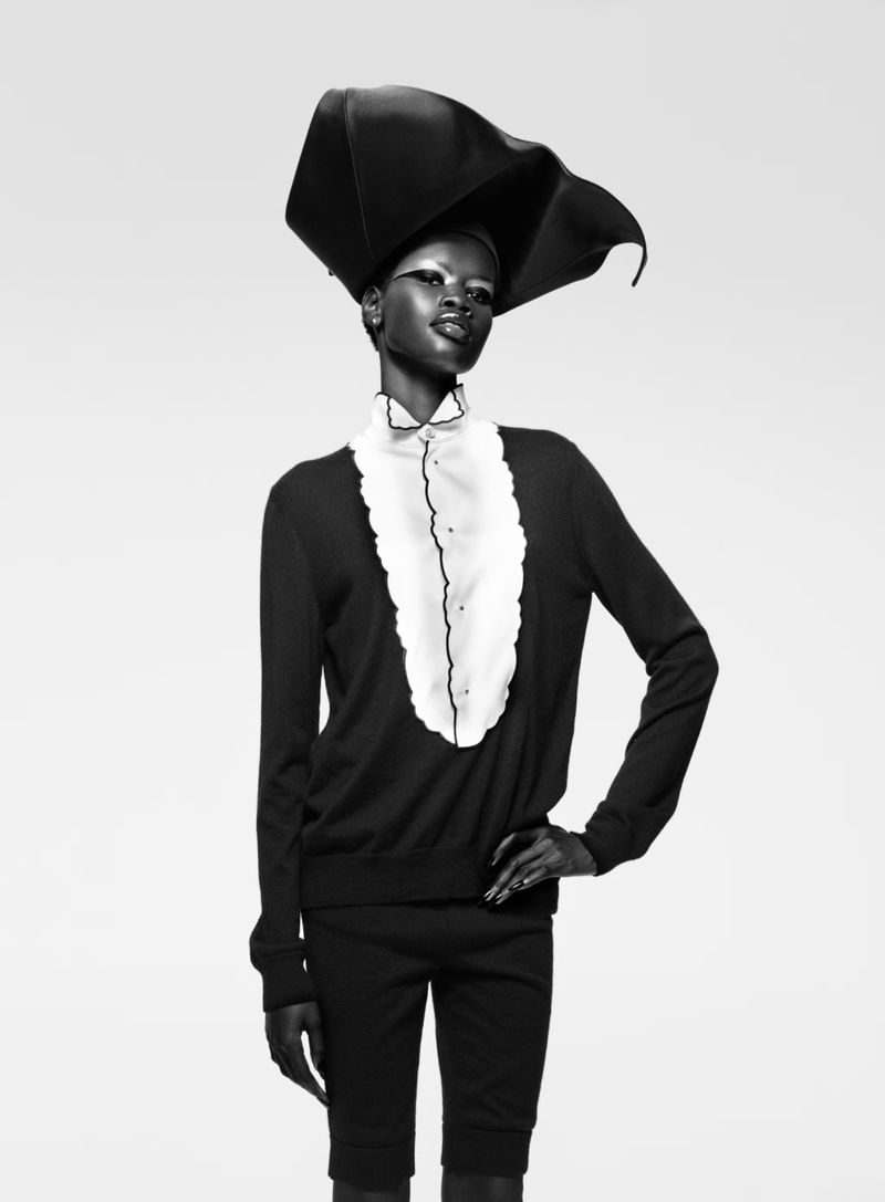 A striking black and white fashion portrait showcases an elegant Black woman modeling a large black hat with a ruffled white collar shirt and dark sweater.