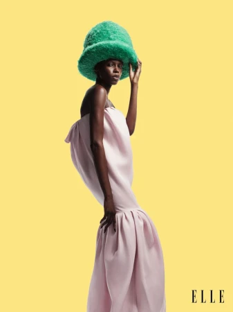 Fashion editorial shot of an African American woman wearing a light pink strapless dress and a large fuzzy green hat against a yellow backdrop.