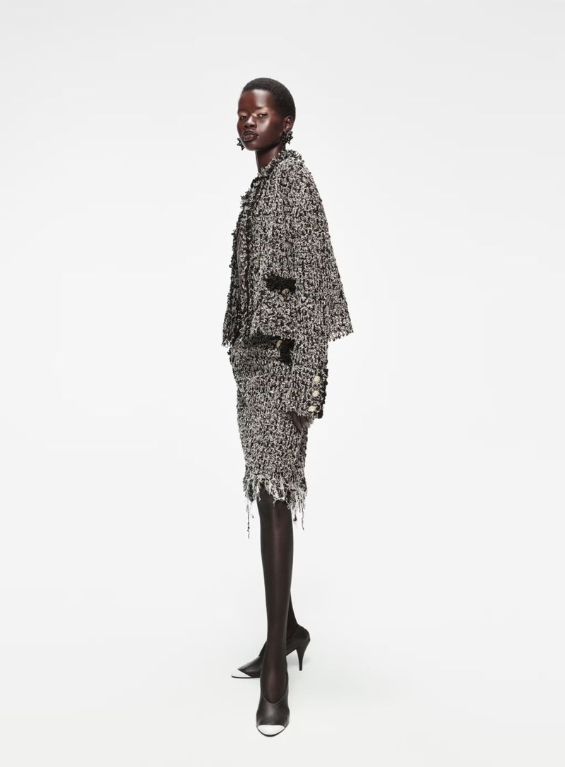 Fashion shot of an African woman posing in a tweed jacket and skirt, standing in a white photo studio in black heels.