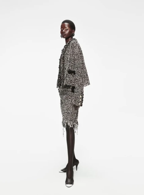 Fashion shot of an African woman posing in a tweed jacket and skirt, standing in a white photo studio in black heels.